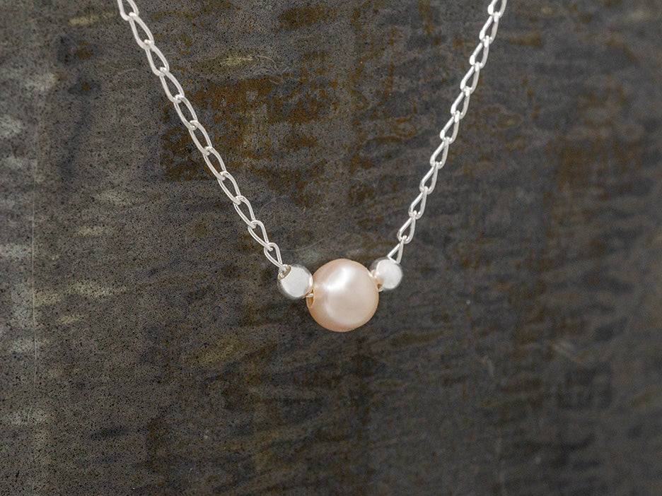 A dainty pink pearl necklace with two silver beads on a silver chain, displayed against a textured background.