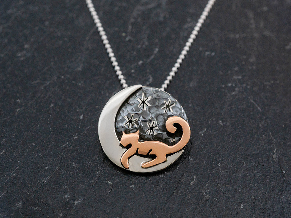 This circular pendant features a copper cat resting on a silver moon against the backdrop of an oxidized sky and silver stars. Pendant hangs on a silver faceted bolitas chain.