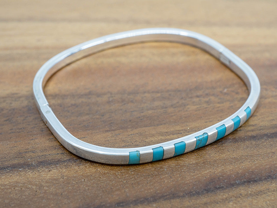 A hinged sterling silver bangle with squares of turquoise inlaid on one side.  The bangle is shaped like a rounded square.