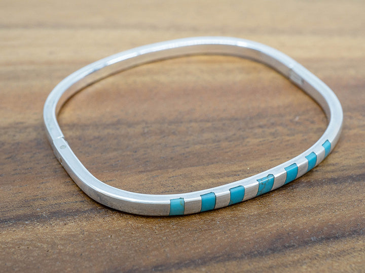 A hinged sterling silver bangle with squares of turquoise inlaid on one side.  The bangle is shaped like a rounded square.