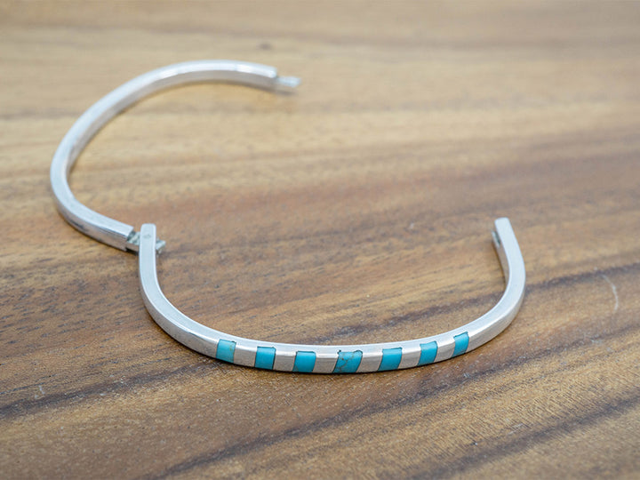 A hinged sterling silver bangle with squares of turquoise inlaid on one side.  The bangle is shaped like a rounded square.