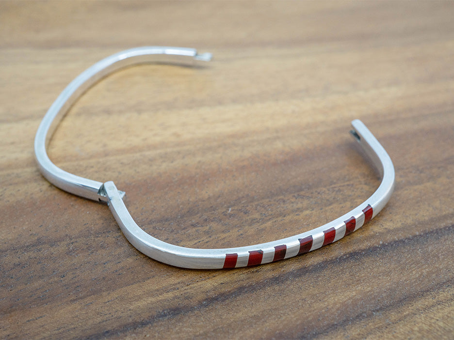 A hinged sterling silver bangle with squares of red jasper inlaid on one side.  The bangle is shaped like a rounded square.