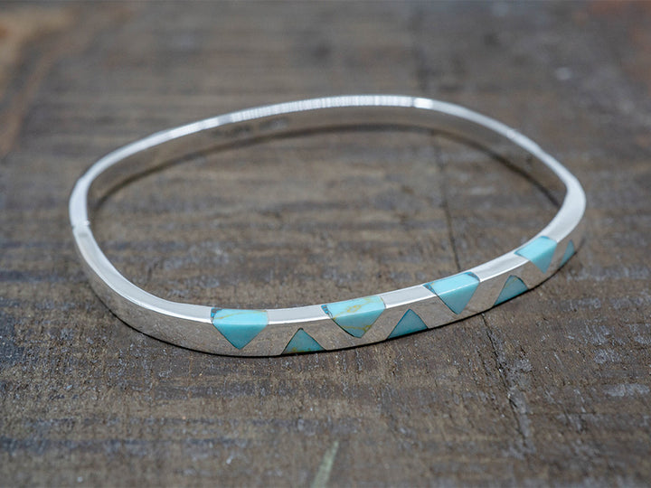A hinged sterling silver bangle with inlaid turquoise triangles on the top. The bangle is shaped like a rounded square.