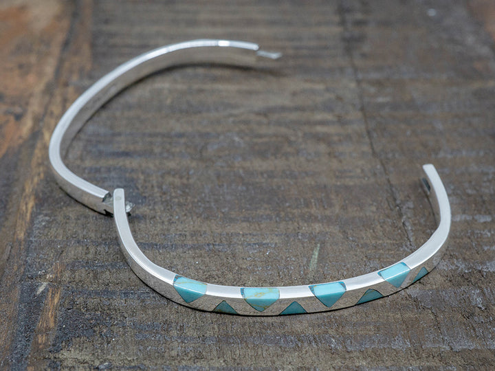 A hinged sterling silver bangle with inlaid turquoise triangles on the top. The bangle is shaped like a rounded square.