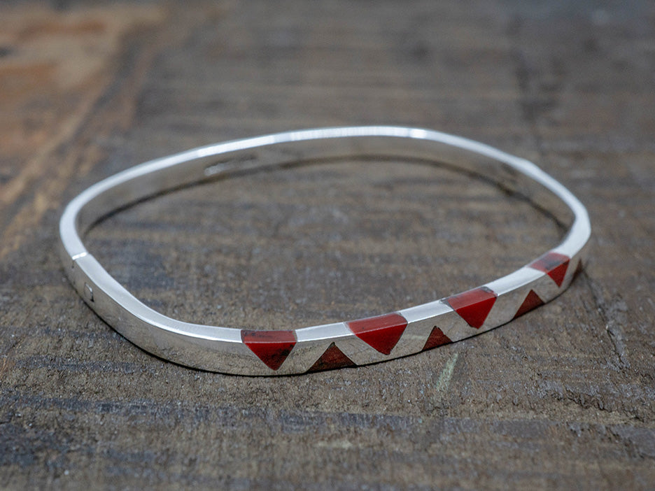 A hinged sterling silver bangle with inlaid red triangles on the top. The bangle is shaped like a rounded square.