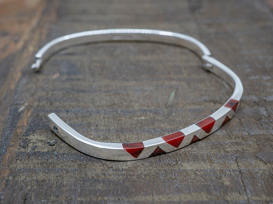 A hinged sterling silver bangle with inlaid red jasper triangles on the top. The bangle is shaped like a rounded square.