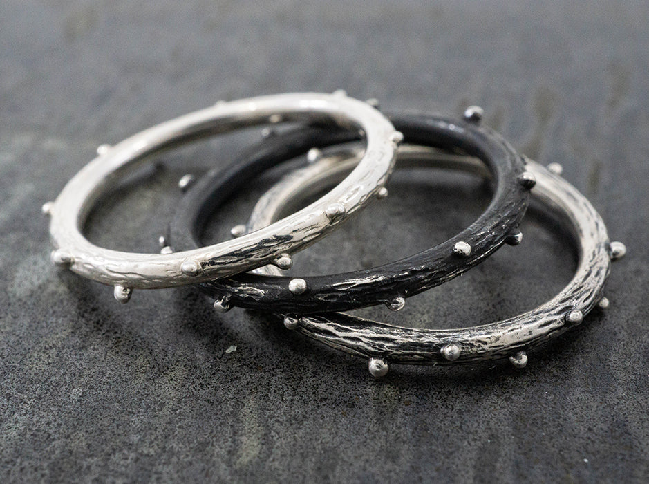 A three piece ring that features one bright, one oxidized and one medium oxidized ring. Each ring has a wood-like texture and silver beads protruding from the surface.