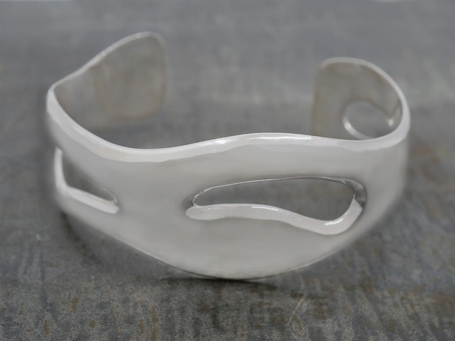 A silver cuff bracelet with a natural, flowing shape displayed on a textured background.