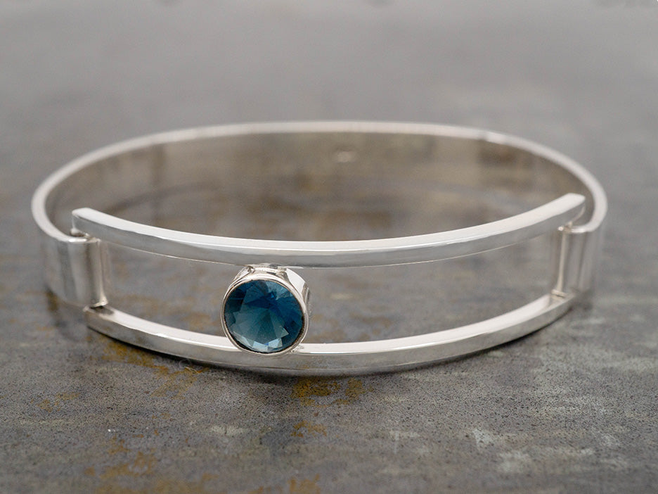 A hinged sterling silver bracelet with an off-center blue crystal in an open-frame.