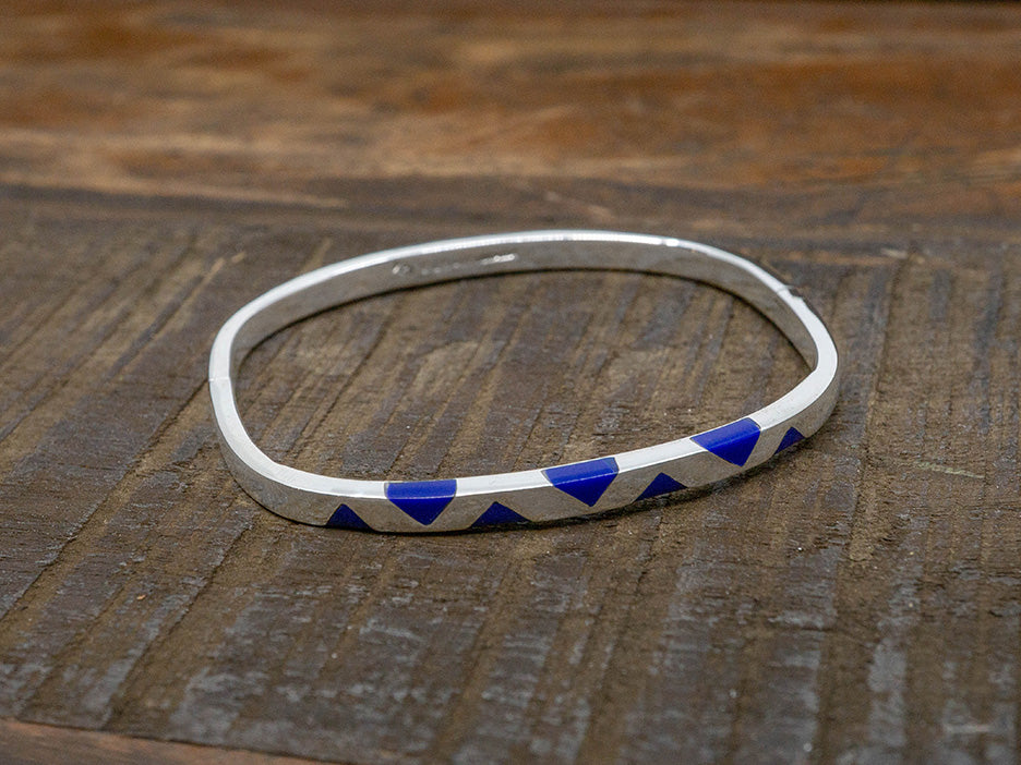 A solid silver bangle in the shape of a rounded square. One side has inlaid lapis-blue triangles.