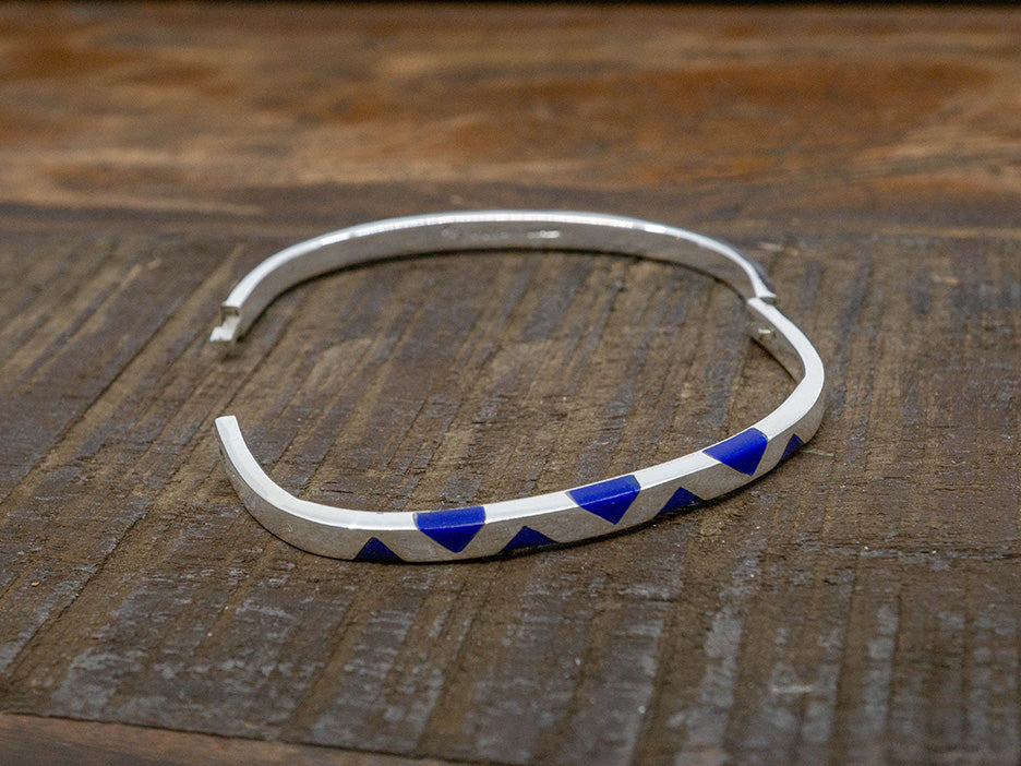 A hinged silver bangle in the shape of a rounded square. One side has inlaid lapis-blue triangles.