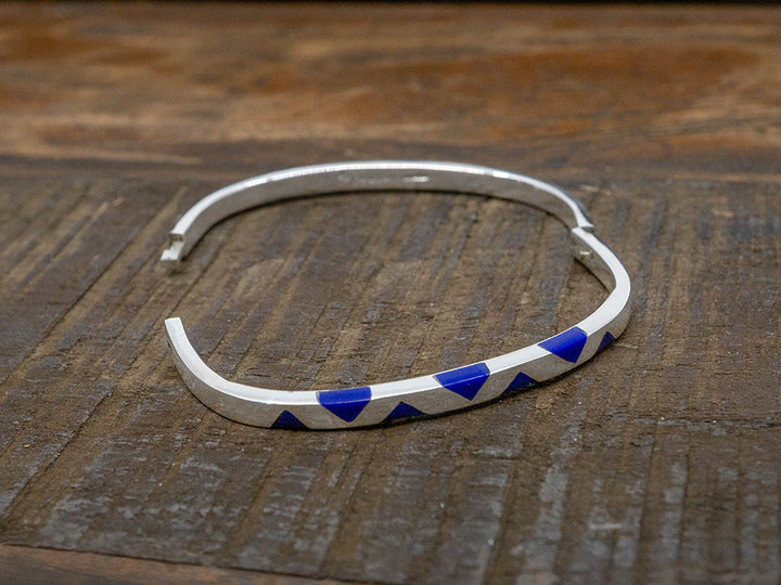 A hinged silver bangle in the shape of a rounded square. One side has inlaid lapis-blue triangles.