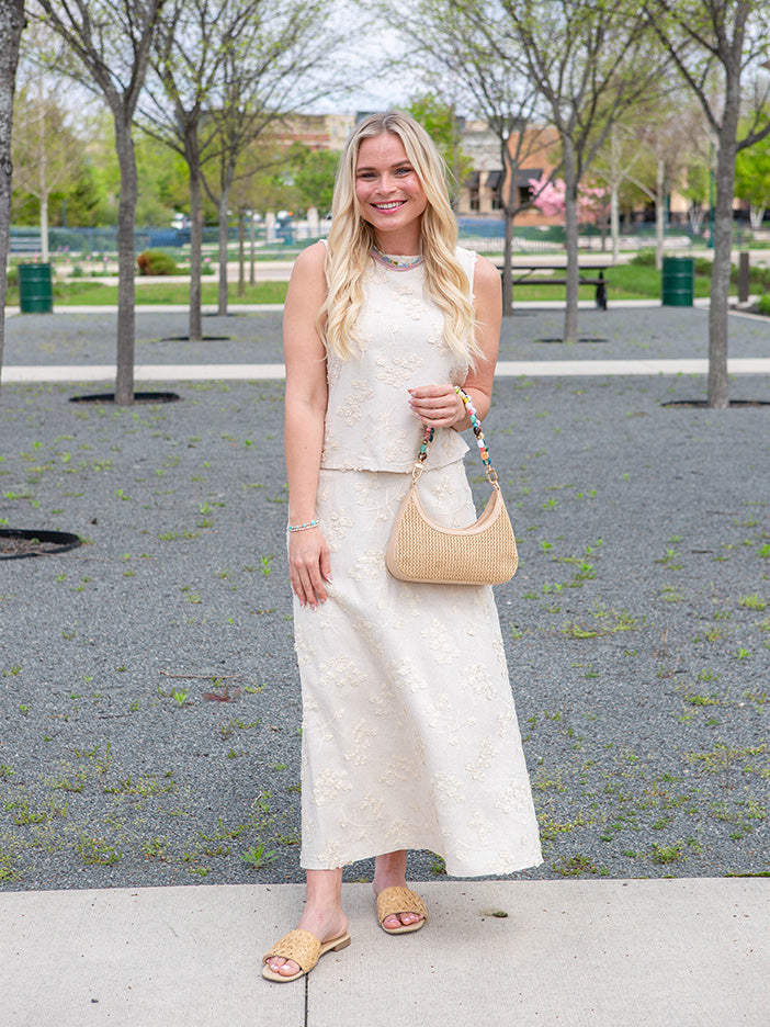 Woman in a white outfit with a beige handbag standing in a park.