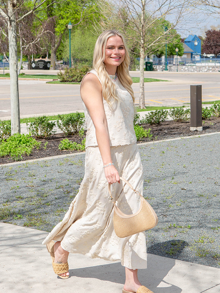 Woman in a light skirt and top holding a beige handbag on a sidewalk with trees and a road in the background.