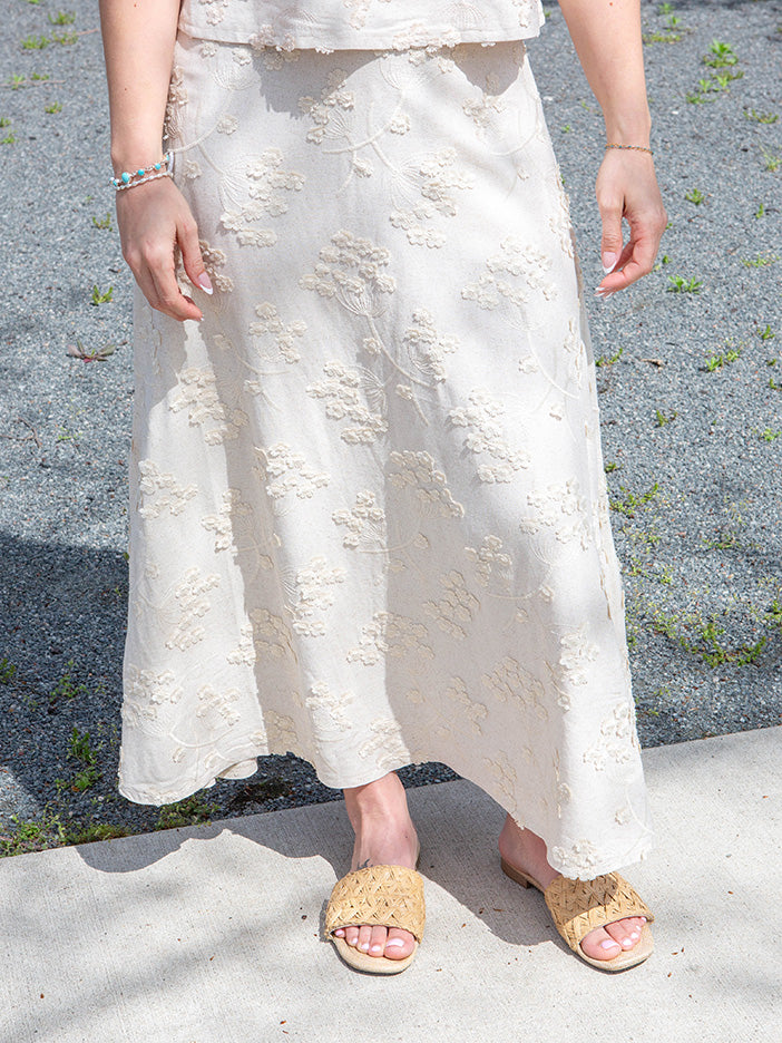 Person wearing a white embroidered skirt with beige sandals on a gray pavement background
