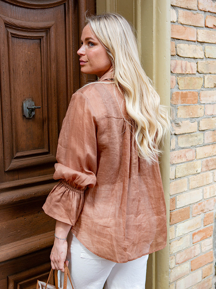 Woman in a brown blouse and white pants standing in front of a wooden door.