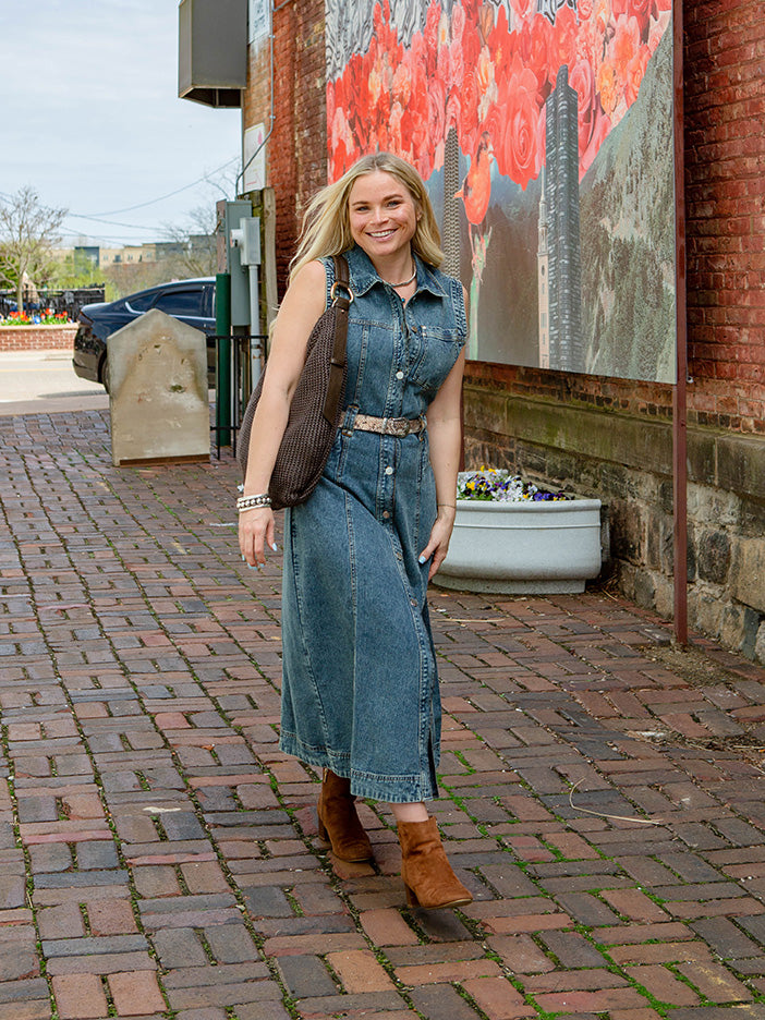 woman walking on cobblestones wearing a denim dress with a large brown handbag