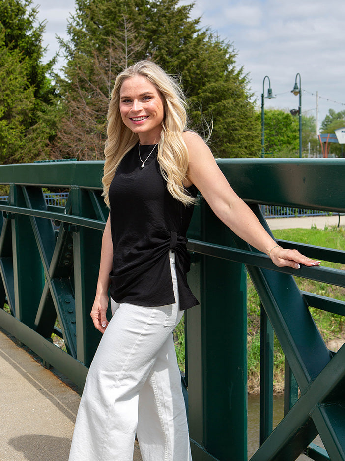 Woman in black top and white pants standing on a bridge with greenery in the background