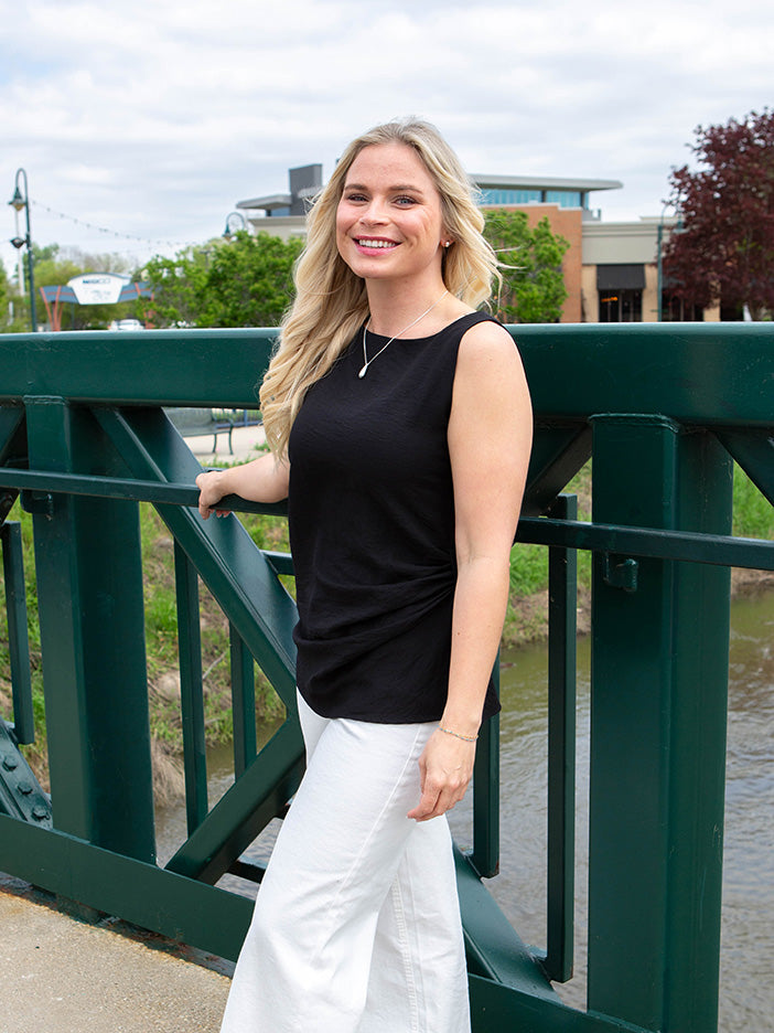 Woman in black sleeveless top and white pants standing on a bridge with a cityscape background
