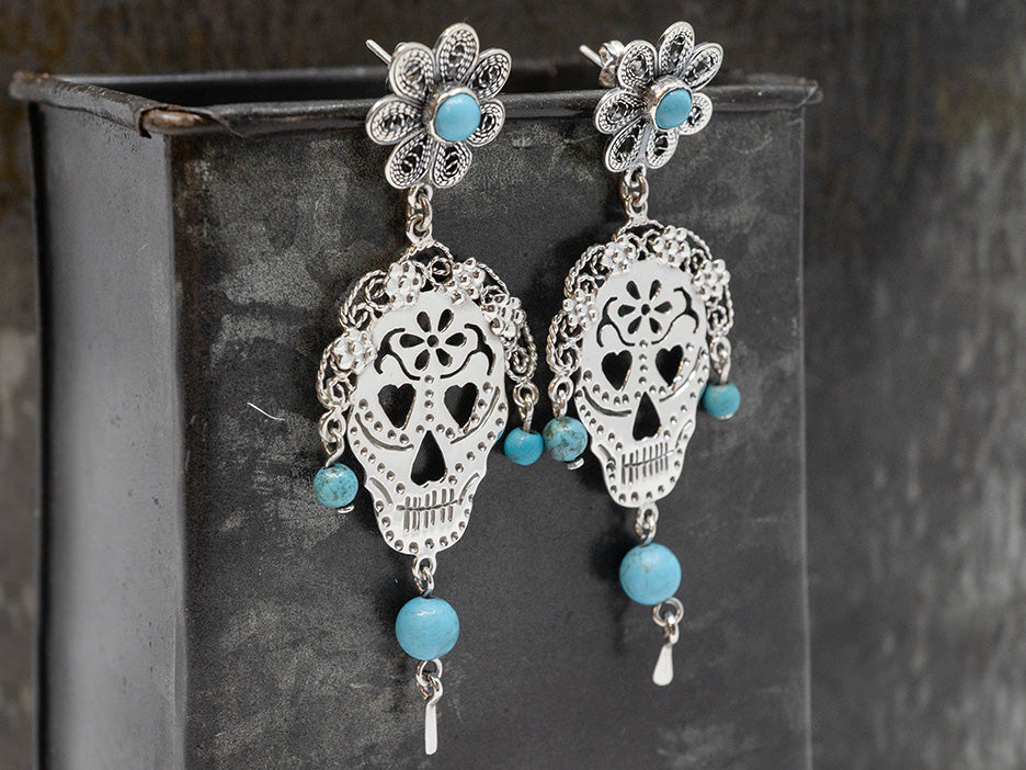 Silver skull-shaped earrings with turquoise beads on a dark background