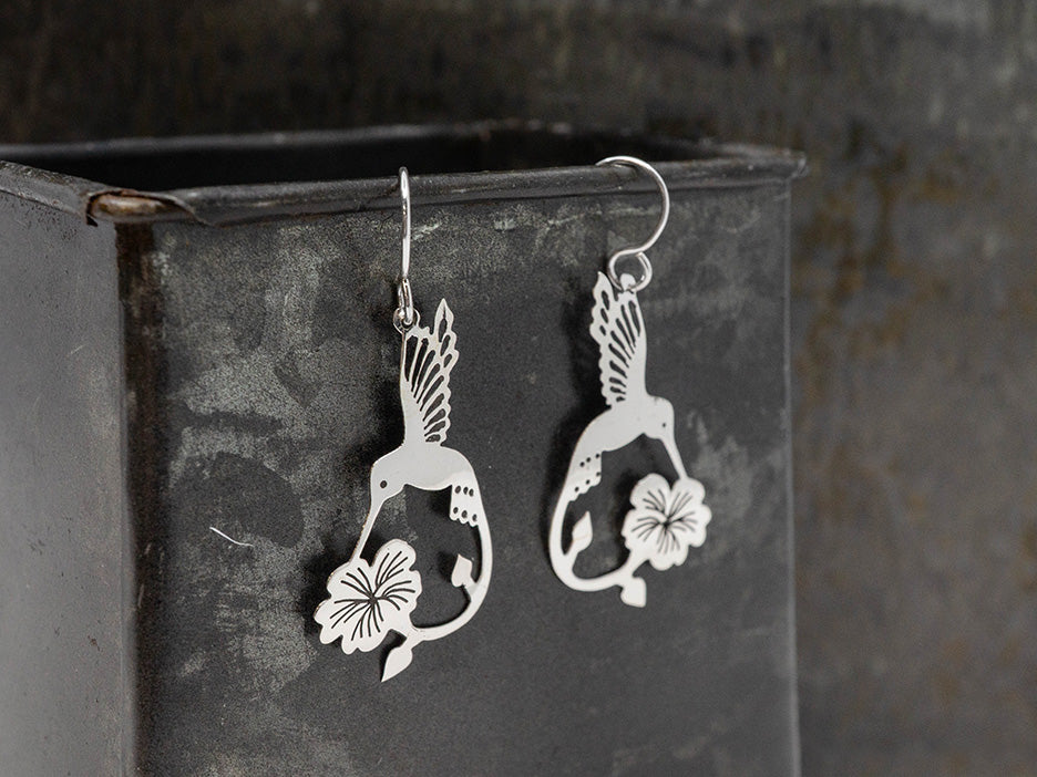 Silver hummingbird earrings with floral designs on a dark metallic surface
