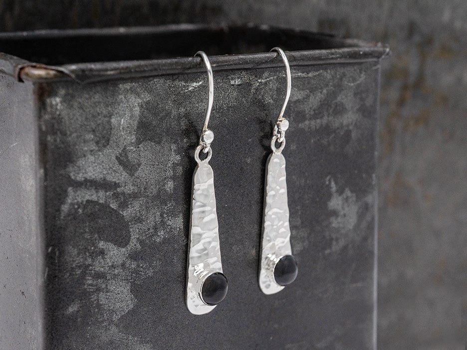 Hammered silver earrings with black accents hanging from a dark metal surface
