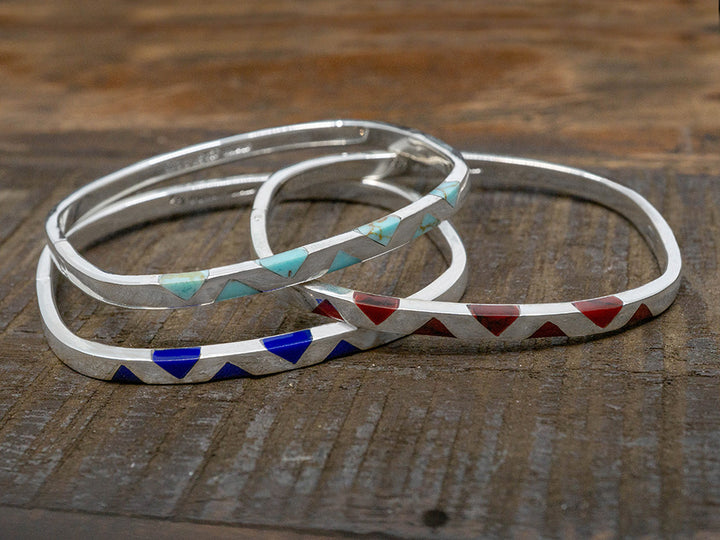 A stack of solid three silver bangles shaped like rounded squares. Each bangle has inlaid stone triangles of different colors.