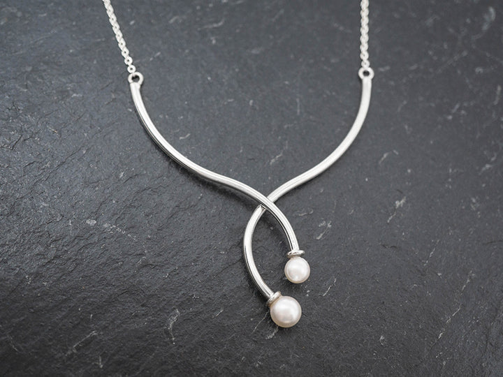 An asymmetrical silver necklace with two crossing tendrils capped with white cultured pearls, displayed against a dark background.