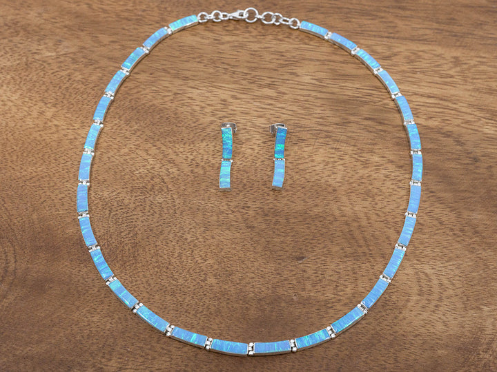 A sterling silver necklace made of rectangular links set with blue opals. A matching pair of earrings sits in the center of the necklace. The earrings each have two links that matching the necklace.