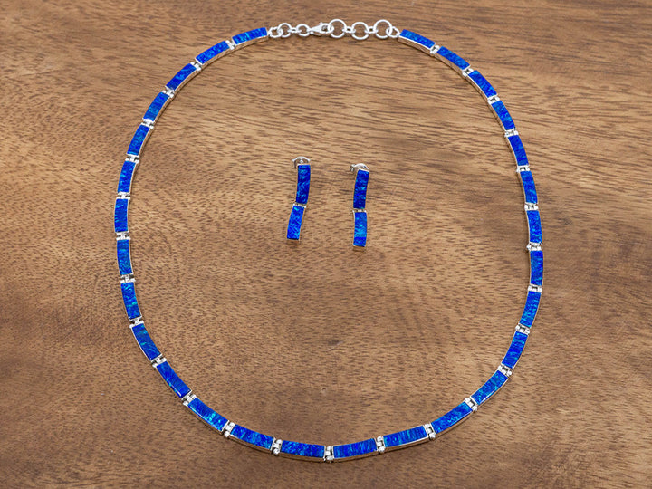 A sterling silver necklace made of hinged rectangular links. Each silver link has dark blue opal set in it. A matching pair of earrings is framed in the center of the necklace. The earrings are each have two links of dark blue opal.