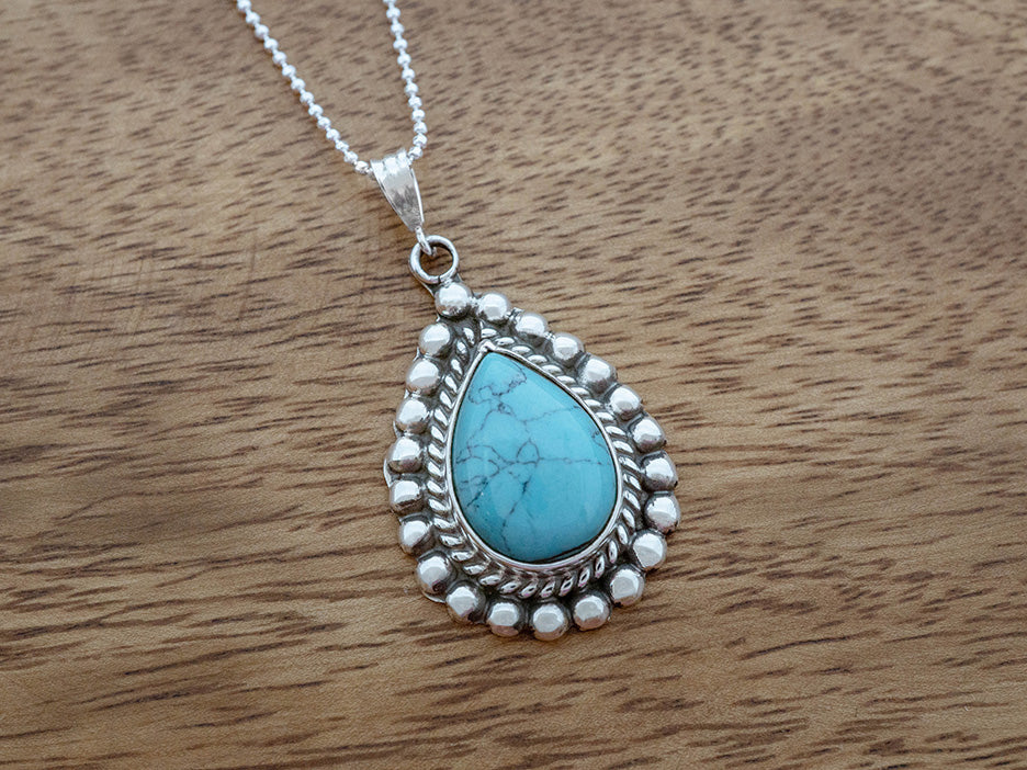 Teardrop-shaped turquoise pendant on a silver chain against a wooden background
