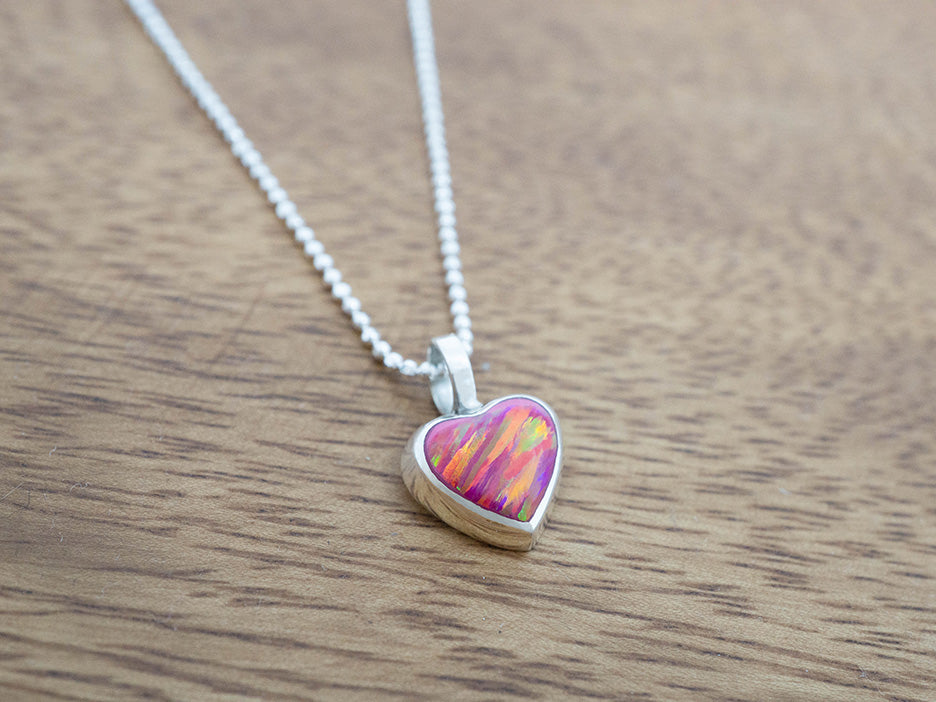Heart-shaped silver pendant with a colorful pink opal center on a silver chain, placed on a wooden surface.