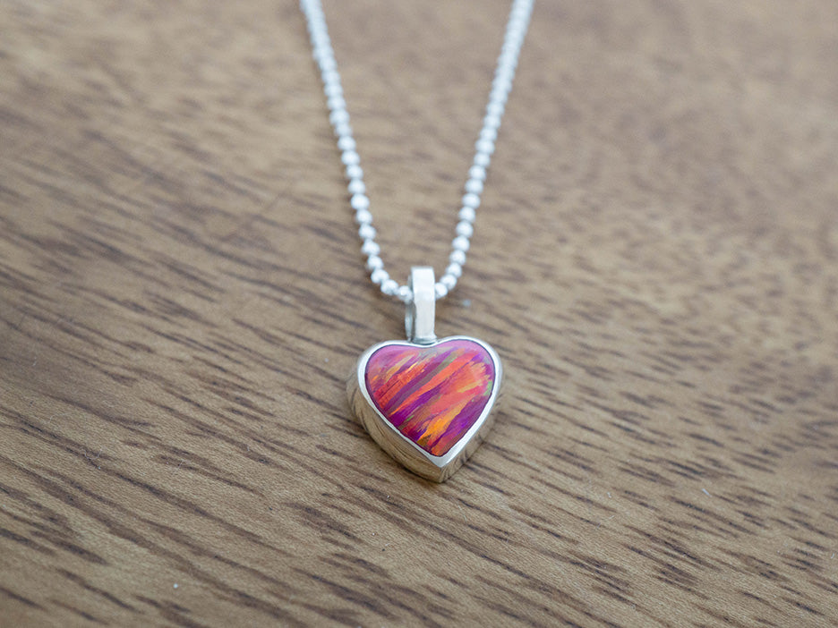 Heart-shaped silver pendant with a colorful pink opal center on a silver chain, placed on a wooden surface.