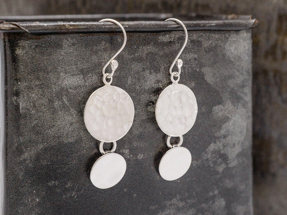 A pair of sterling silver earrings. Each earring features a smooth silver disc suspended from a larger, hammered silver disc. 